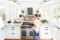 Bright kitchen with white cabinets, large island, modern pendant lights, and a person carrying plates.