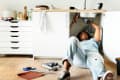 Person fixing a kitchen sink, lying on the floor with tools and pipes nearby, next to white cabinets.