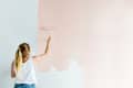 Person painting a wall light pink with a roller, wearing a white tank top and jeans.