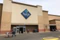 The exterior of a Sam’s Club store with a shopper walking in and away from it.