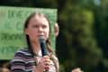 Young person with braided hair speaking into a microphone outdoors, holding a green protest sign in the background.
