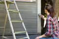 Woman worker painting wooden house exterior wall with paintbrush and wood protective color home improvement concept.