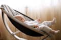 Cute smiling baby laying in bouncer chair. Child relaxing in a swing. Adorable newborn baby in bodysuit in sunny bedroom. Family morning at home.