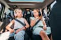 Portrait of two positive smiling sisters sitting in child car seats fastening safety belts listening to music using headphones. Happy childhood, auto journey concept image.