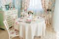 Enchanting vintage dining room with delicate floral patterns, pastel green furniture, and elegant table set with books and flowers. Sunbeams shine through sheer curtains, creating soft, inviting glow