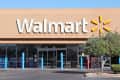 Walmart storefront with large logo, glass entrance, blue bollards, and a tree on the right.