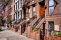 Brownstone row houses with ornate iron railings and steps, potted plants, and a tree-lined sidewalk.