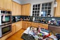 Cluttered kitchen with wooden cabinets, stainless steel appliances, and a messy countertop filled with various items.