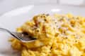 Creamy scrambled eggs topped with grated cheese and black pepper on a white plate, with a fork lifting a portion.