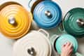 Colorful cast iron Dutch ovens in yellow, blue, green, and cream, with a hand lifting the lid of the teal pot.