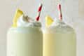 Two creamy lemon drinks topped with whipped cream and lemon slices, each with a red and white striped straw.