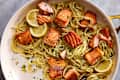 Pasta with sautéed salmon pieces, garnished with lemon slices and herbs in a white skillet.
