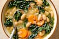 Bowl of soup with white beans, kale, carrots, and spices, served with a spoon.