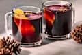 Two clear mugs filled with dark red beverage, garnished with orange peels and spices, beside pinecones.