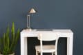 White desk with a closed laptop, silver lamp, and white chair against a dark blue wall, next to a tall green plant.