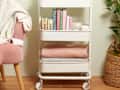 living room vignette with a white rolling cart being used to organize blankets, books, and art supplies between a chair with a blanket draped over it and a plant