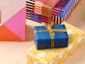 Colorful wrapped gifts stacked together, featuring a blue box with a gold ribbon on a yellow patterned box.