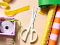 Crafting supplies on a table featuring scissors, colorful ribbons, and patterned wrapping paper rolls.