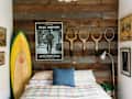 A surfboard, posters, and tennis rackets above a bed in a rustic bedroom.