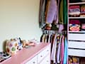 Pink dresser with decorative jars and trinkets beside a closet with colorful clothes on hangers and shelves.