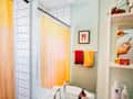 bathroom with shower with white tiles with black grout, pale orange shower curtain, pale sage green walls, shelves with grooming products, reed diffuser, white pedestal sink, red and pale orange accents