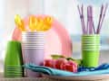 Stacked colorful plastic cups, plates, and utensils with pink tulips and blue napkins on a table.