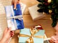 Overhead shot of someone handing over a gift wrapped in silver and blue wrapping paper, and tied in a silver bow, over a light wood surface.  On the light wood surface theres an assortment of other wrapped gifts and gift bags.