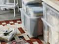 Clear storage bins filled with folded clothes under a bed, with a white step stool and a patterned rug nearby.