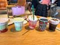 Five colorful drinks on a wooden table, with pastries and a pink handbag in the background.