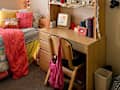 Cozy bedroom with a colorful bedspread, decorative pillows, a wooden desk with two chairs, and a shelf filled with books and decor.