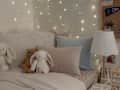 Cozy bedroom with fairy lights, plush toys, pastel bedding, and a bedside lamp on a wooden nightstand.