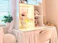 Vanity desk with a mirror and lights, pink flowers, decorative items, and a cozy chair, near a window with white curtains.