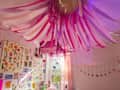 Colorful ceiling decorations with pink and white streamers, floral wall art, and a window with sheer curtains.