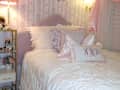 Cozy bedroom with a lavender headboard, ruffled white bedding, decorative pillows, and a bedside table with a lamp.