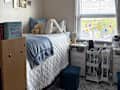 Cozy bedroom with a bed covered in a white quilt, blue throw, and decorative pillows, featuring a stuffed bear, string lights, and a colorful banner.