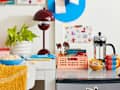 closeup shot of desk that has a red lamp, white planter, supplies and a blue bowl of popcorn. next to the desk is a mini fridge with a mug, coffee press, and a pink basket with utensils coffee assortments on it