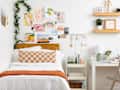 forward shot of bedroom where the bed has orange and cream checkered pillow and orange blanket, a gallery wall above the headboard - a rolling cart next to bed and a desk
