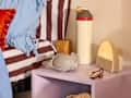 A bedside table with a lavender finish, featuring headphones, a water bottle, a wooden block, and a lamp.