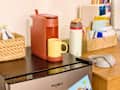 Keurig coffee maker in terracotta color with a yellow mug, a water bottle, and a basket of snacks on a wooden table.