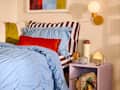 Cozy bedroom corner featuring a blue ruffled duvet, striped pillows, an orange velvet accent pillow, and a purple nightstand with decor.