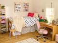 Cozy bedroom with a lofted bed, polka dot bedding, a pink desk chair, and colorful decor items on a wooden desk.