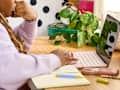 A person with long hair in a lavender sweatshirt is using a pink laptop on a wooden desk, surrounded by a notebook, pens, and a green plant.