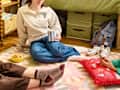 Two young women sitting on a colorful rug, one holding a striped mug, with snacks and a red decorative pillow nearby.