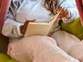A person wearing a gray sweatshirt and pink pants sits on a green cushion, writing in a notebook with a pink pen.