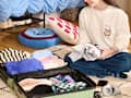 A person sitting on the floor, packing a suitcase with colorful clothes, surrounded by decorative pillows and a phone.