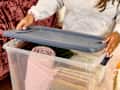 A person in a gray sweatshirt opening a clear storage bin filled with rolled blankets and books.