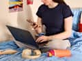 A person sitting on a bed with a blue comforter, using a laptop and holding a phone, with headphones and a colorful container nearby.
