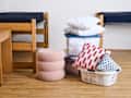 A stack of pillows and colorful cushions next to a laundry basket in a dorm room with wooden furniture.