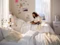 A cozy bedroom with two beds, a woman reading on one, soft blankets, a small table, and a mini fridge.