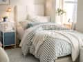 Cozy bedroom with a striped duvet, white headboard, bedside table, lamp, and a plant by the window.